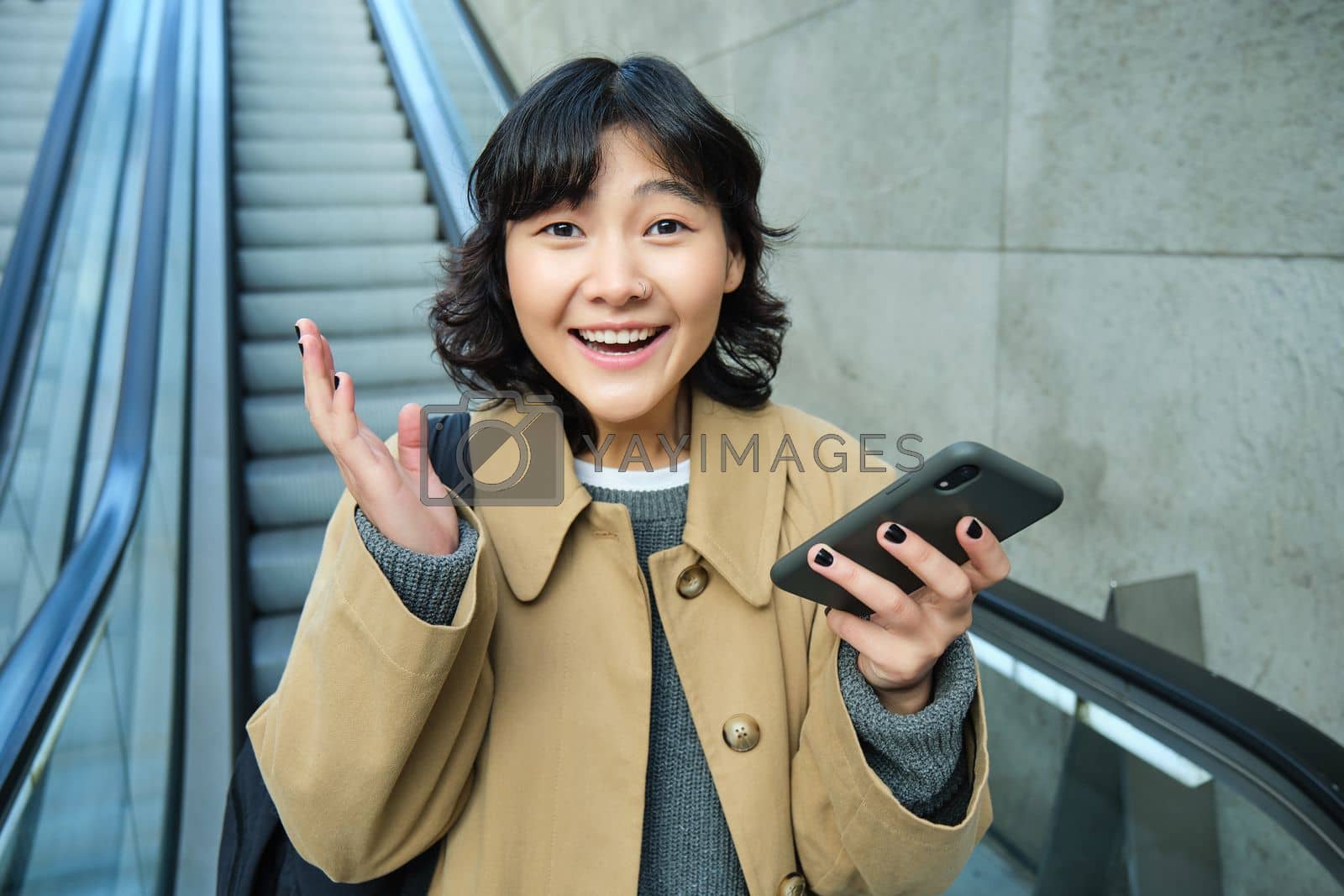 Royalty free image of Happy triumphing asian girl, looks amazed and joyful, holds smartphone, goes down escalator with pleased face, wins, hears good news by Benzoix