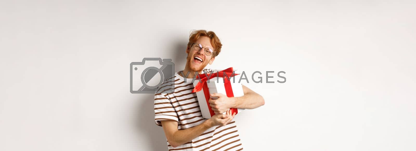 Royalty free image of Valentines day and holidays concept. Happy redhead man hugging his gift box and thanking, looking at camera excited and grateful, white background by Benzoix