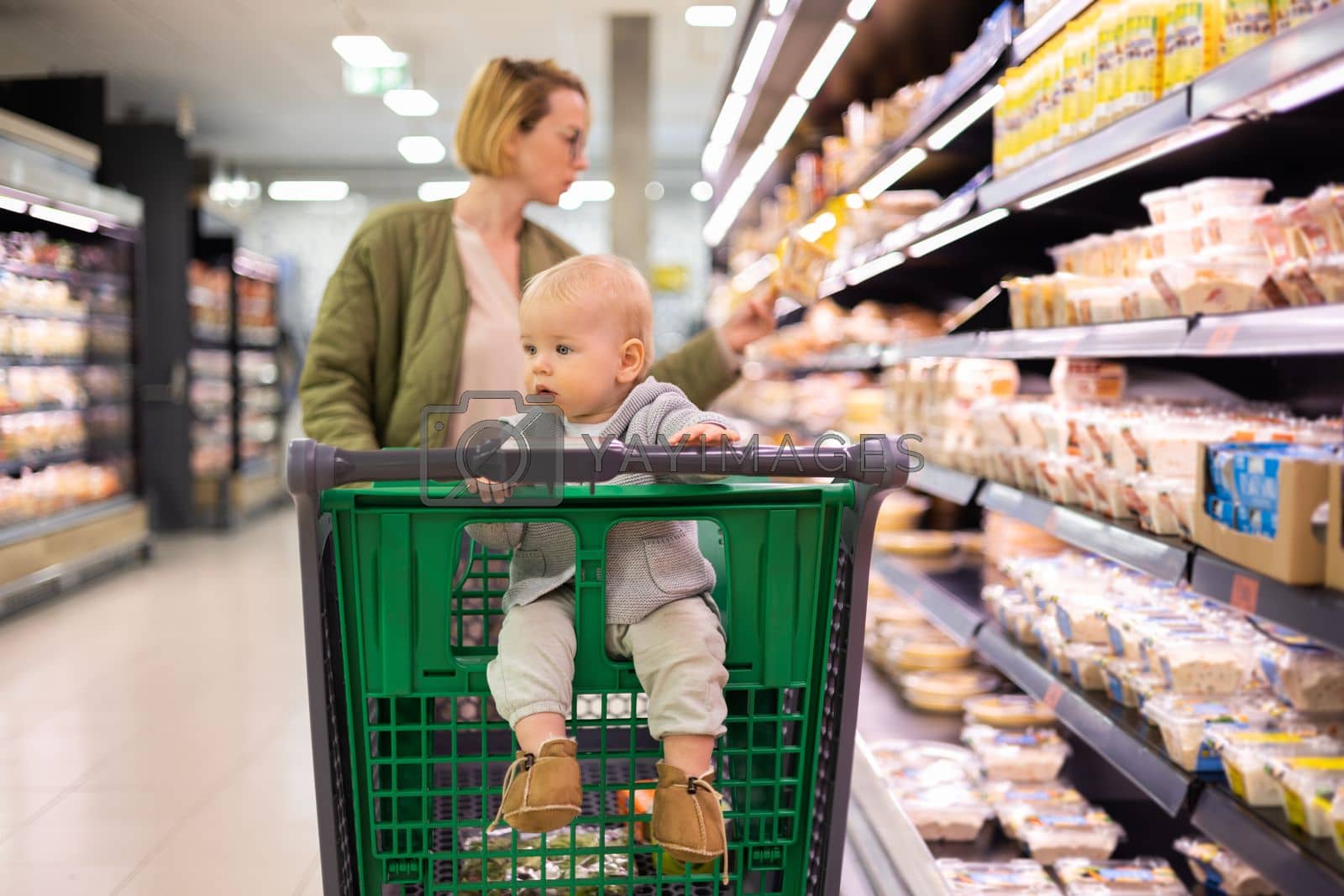 Royalty free image of Mother pushing shopping cart with her infant baby boy child down department aisle in supermarket grocery store. Shopping with kids concept. by kasto