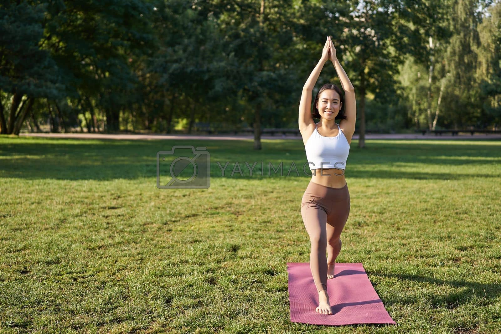 Royalty free image of Vertical shot of asian woman standing in tree pose, making yoga asana on fresh air, wellbeing training in park on rubber mat by Benzoix