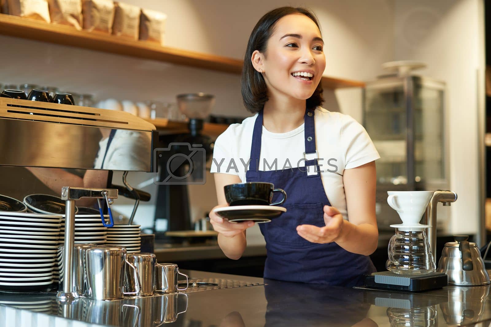 Royalty free image of Portrait of beautiful asian girl, student working part-time in cafe, holding cup of coffee, made order, looking for client, wearing uniform by Benzoix