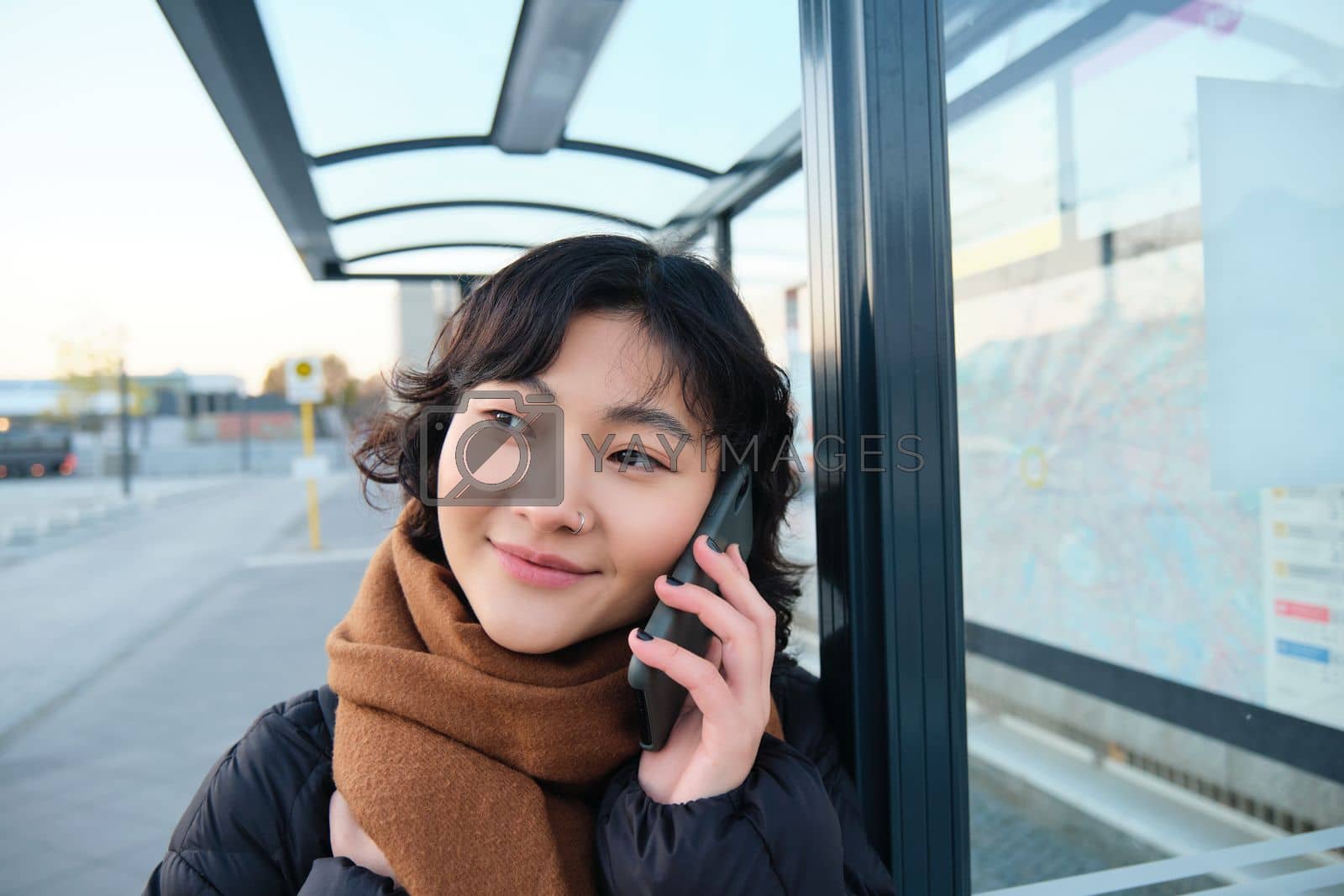 Royalty free image of Close up portrait of cute young woman calling someone, waiting on bus stop with smartphone, using telephone while expecting public transport to arrive by Benzoix