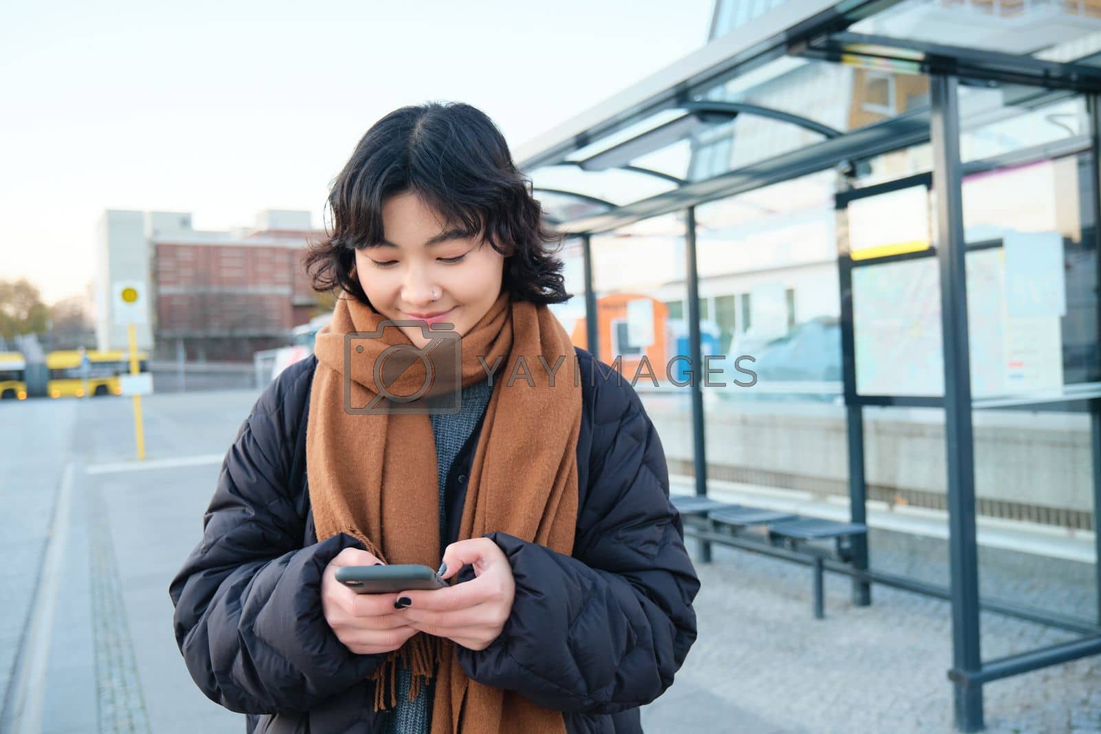 Royalty free image of Happy smiling korean girl, using mobile phone, standing on bus stop with smartphone, looking at departure schedule on application, posing in winter clothes by Benzoix