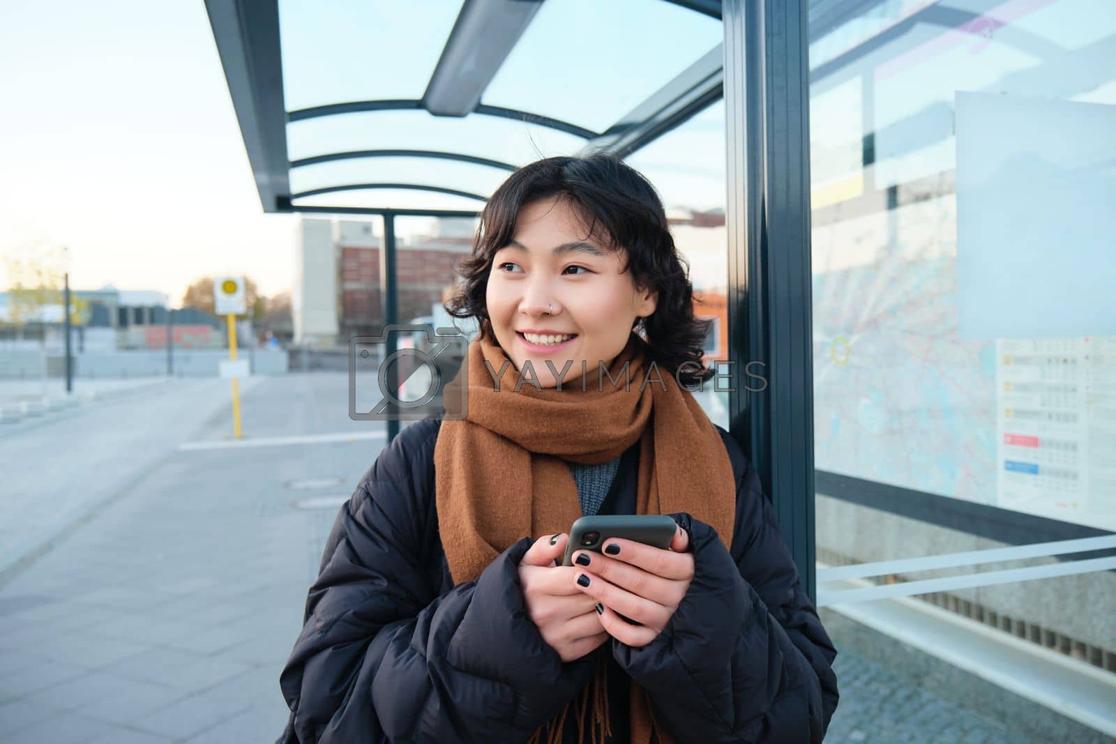 Royalty free image of Portrait of Korean woman in winter jacket, standing with smartphone, waiting for bus on stop, looking at mobile phone app checking public transport application by Benzoix