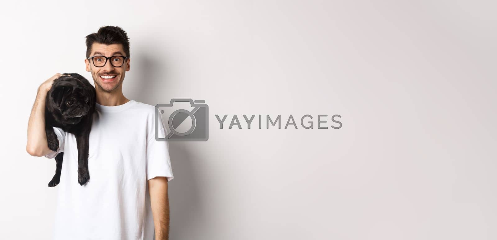 Royalty free image of Cheerful hipster man in glasses, holding cute black pug dog on shoulder, staring at camera with happy smile, standing over white background by Benzoix