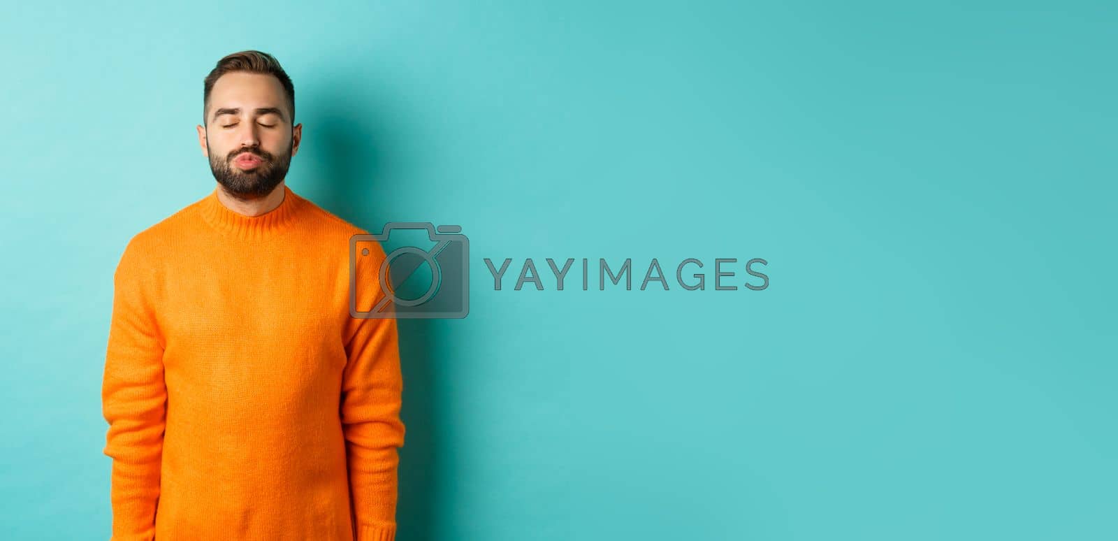 Royalty free image of Waist up shot of young man pucker lips and close eyes, waiting for kiss, standing in orange sweater against light blue background by Benzoix