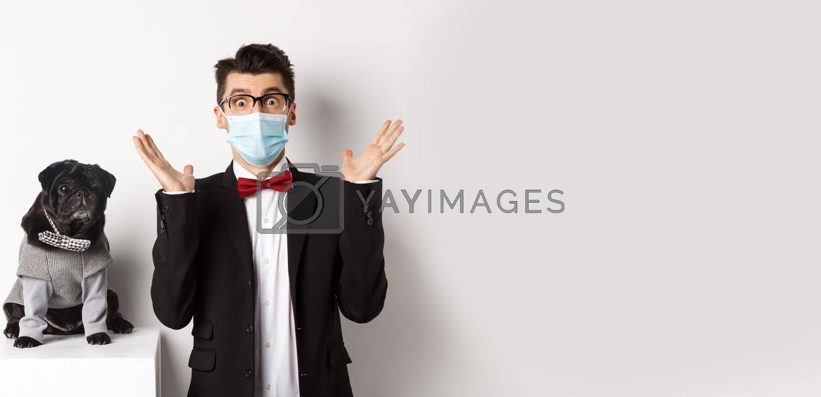 Royalty free image of Coronavirus, pets and celebration concept. Amazed young man in face mask and suit staring at camera surprised, cute black dog sitting near owner in party outfit, white background by Benzoix