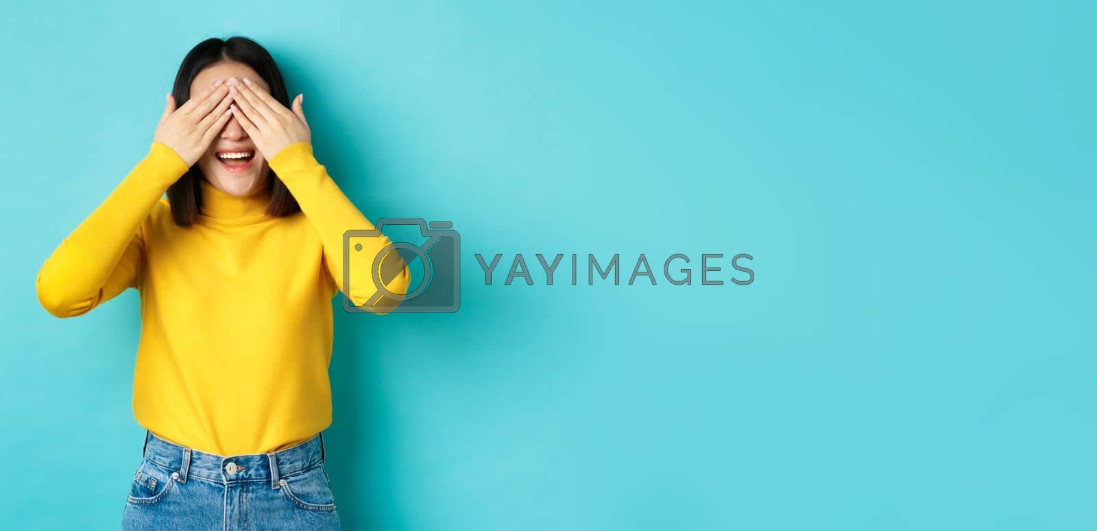 Royalty free image of Cheerful asian girl in yellow pullover waiting for surprise, playing hide n seek and smiling, expecting gift with eyes closed, standing over blue background by Benzoix