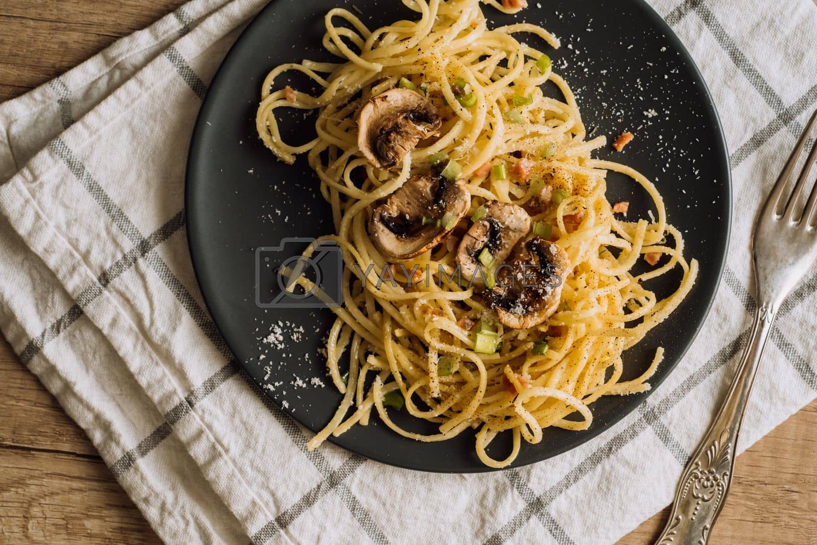 Royalty free image of Spaghetti pasta with champignon mushrooms sprinkled with cheese parmesan and green onions on a black plate with a towel and a fork on a wooden table by Romvy