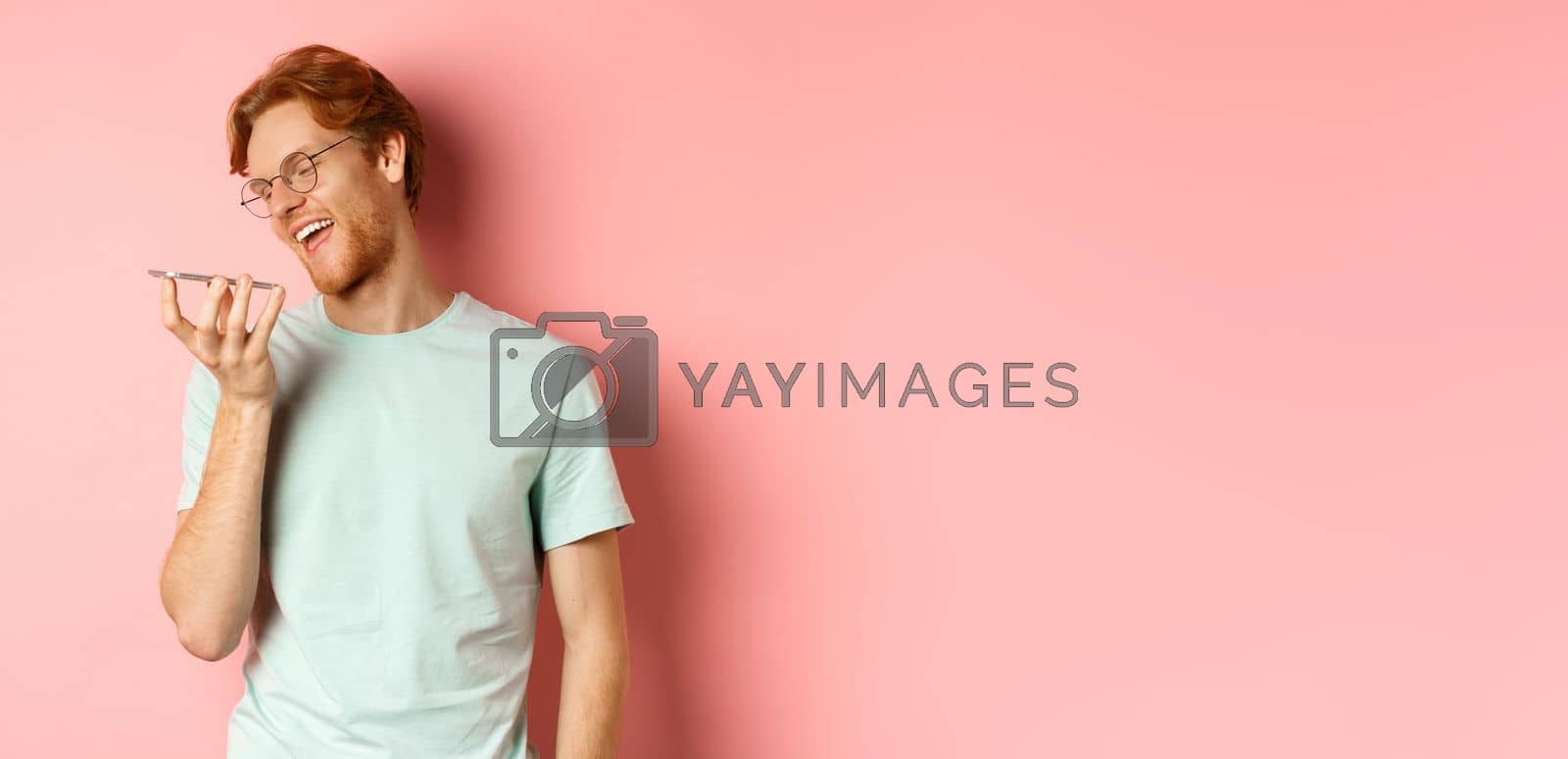 Royalty free image of Happy young man with red hair, smiling pleased while record voice message on smartphone, talking to virtual assistant, standing over pink background by Benzoix