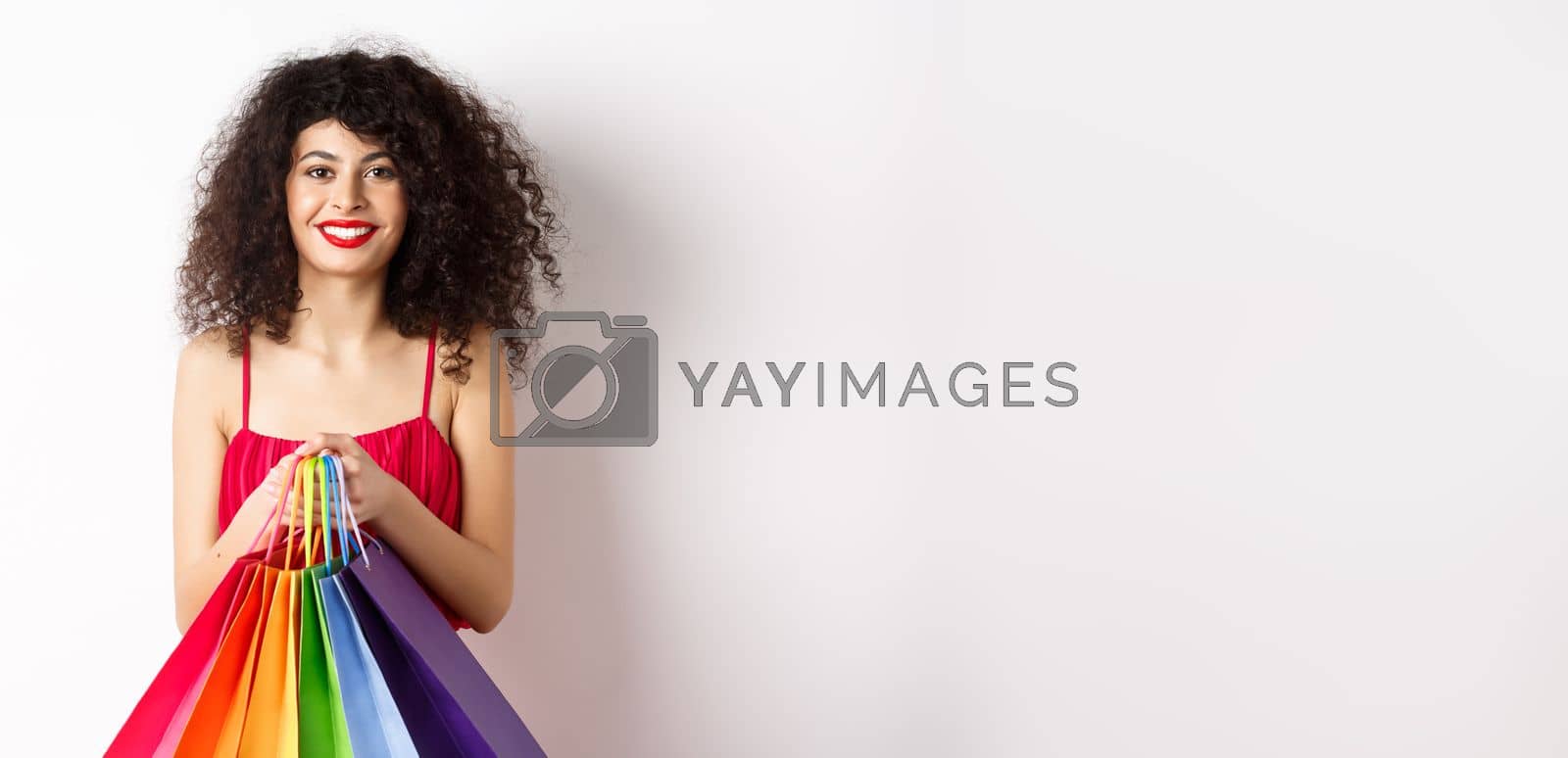 Royalty free image of Image of stylish caucasian woman in red dress and makeup, holding shopping bags and smiling, standing over white background by Benzoix