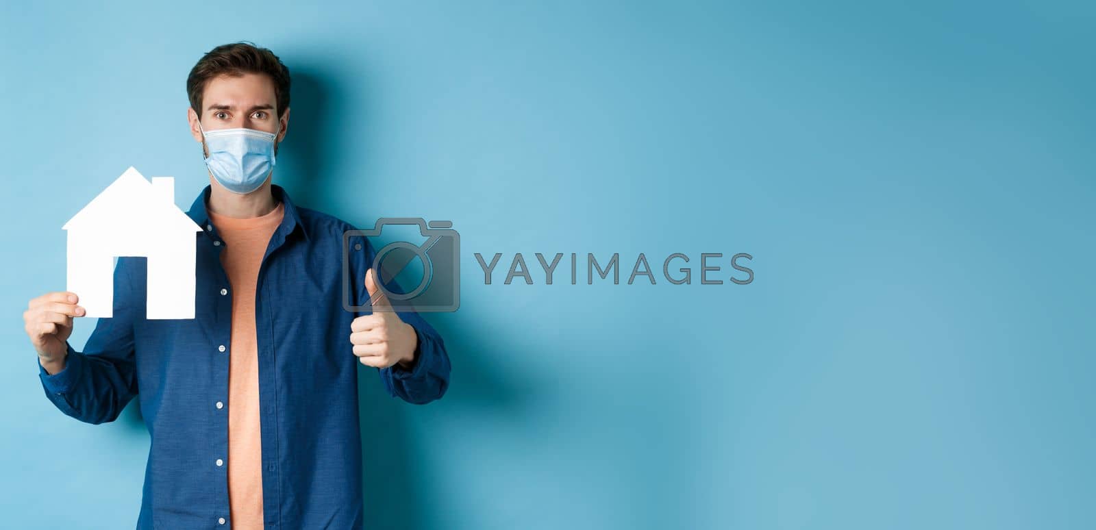 Royalty free image of Real estate and covid concept. Young man in face mask showing paper house cutout and thumb-up, recommending agency, standing on blue background by Benzoix