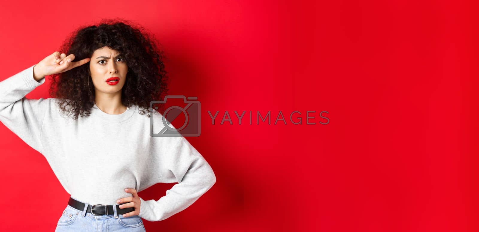 Royalty free image of Are you stupid. Annoyed and confused girl with curly hair, pointing at head and scolding person being crazy or strange, standing on red background by Benzoix