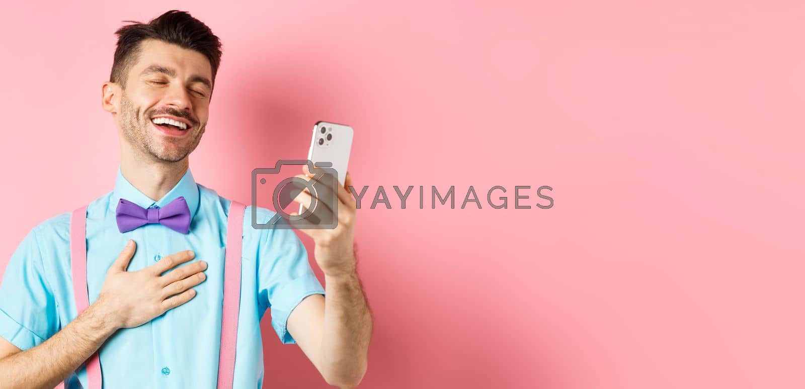 Royalty free image of Technology concept. Happy caucasian man video chat and laugh at smartphone, standing over pink background by Benzoix