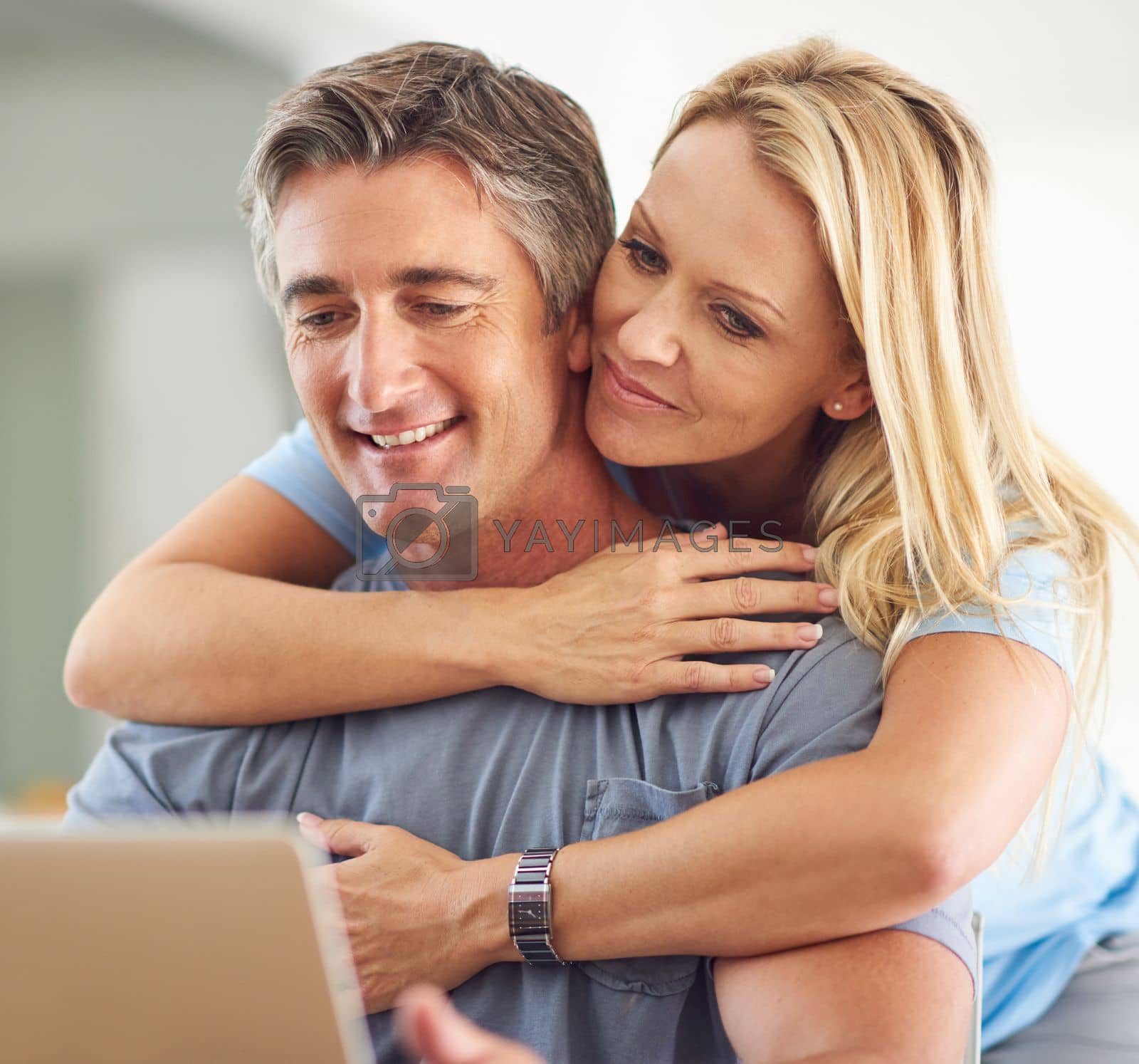 Royalty free image of He completes me. an attractive mature woman hugging her handsome mature husband while he uses a laptop in the house. by YuriArcurs