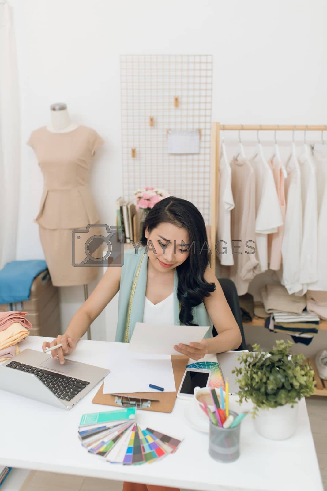 Royalty free image of Fashion designer at work. Happy young Asian woman drawing while sitting at her working place in fashion workshop by makidotvn