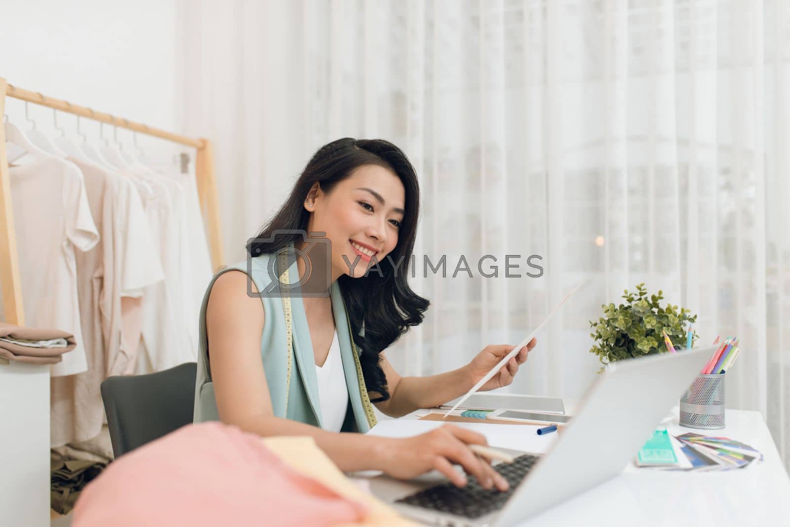 Royalty free image of Fashion designer at work. Happy young Asian woman drawing while sitting at her working place in fashion workshop by makidotvn