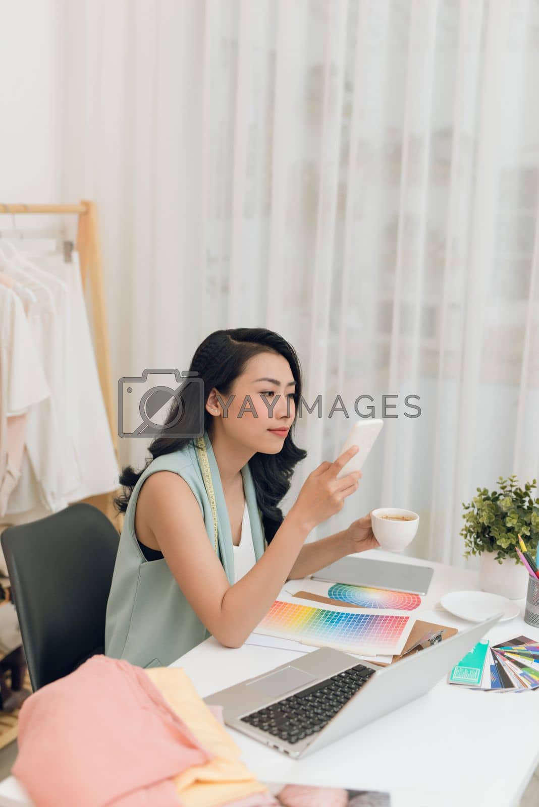 Royalty free image of Fashion designer using smartphone and drinking coffee in her office by makidotvn