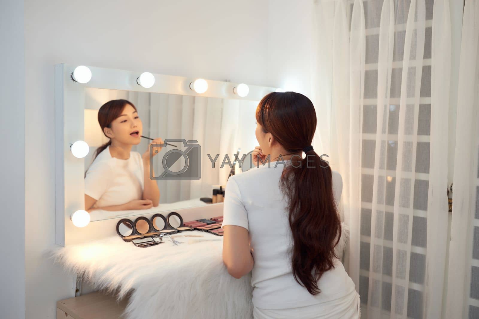 Royalty free image of Beautiful Asian girl applying lipstick watching in mirror at makeup table by makidotvn
