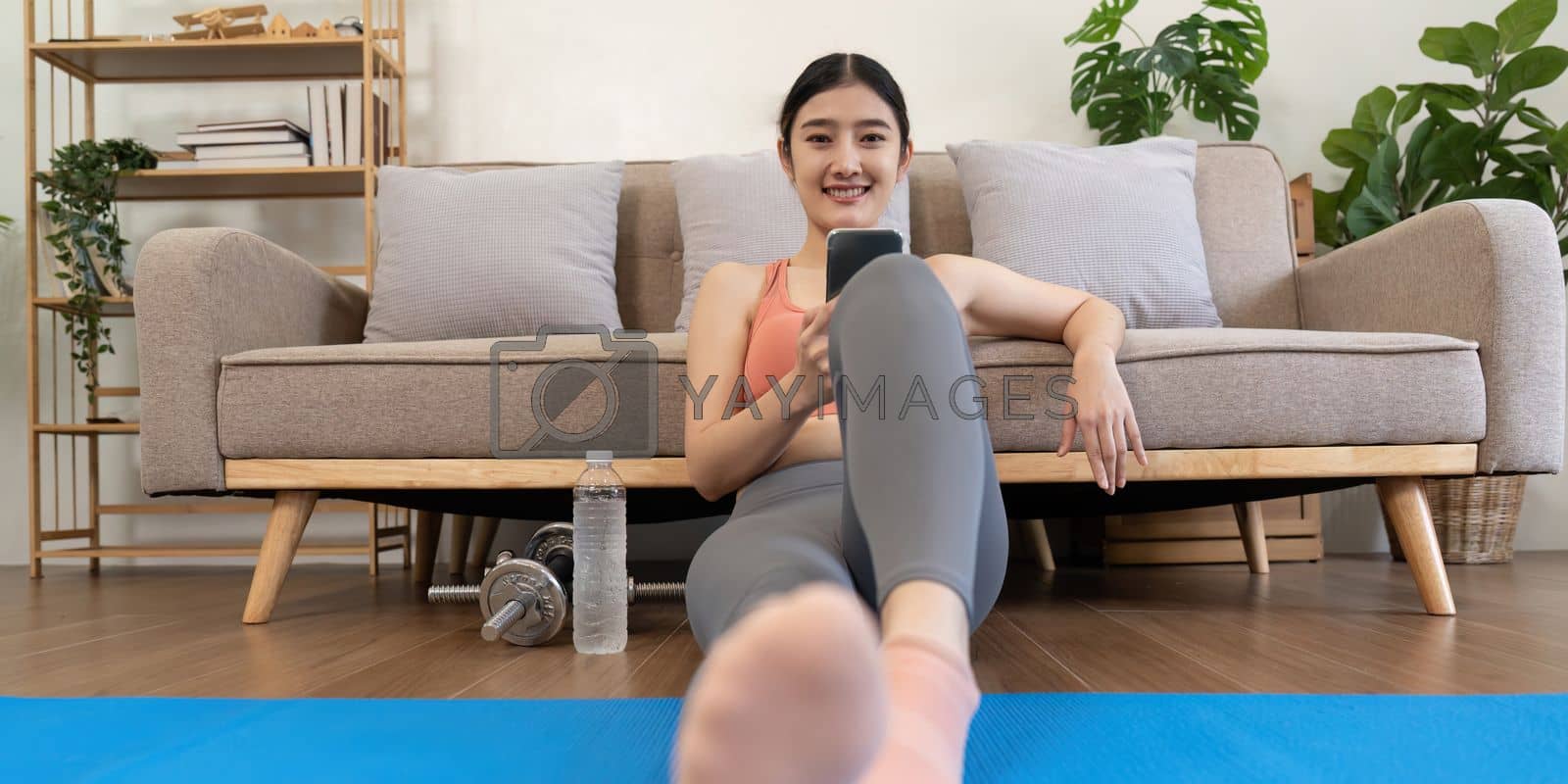Royalty free image of young woman exercising at home happily sitting and resting playing on the mobile phone by nateemee