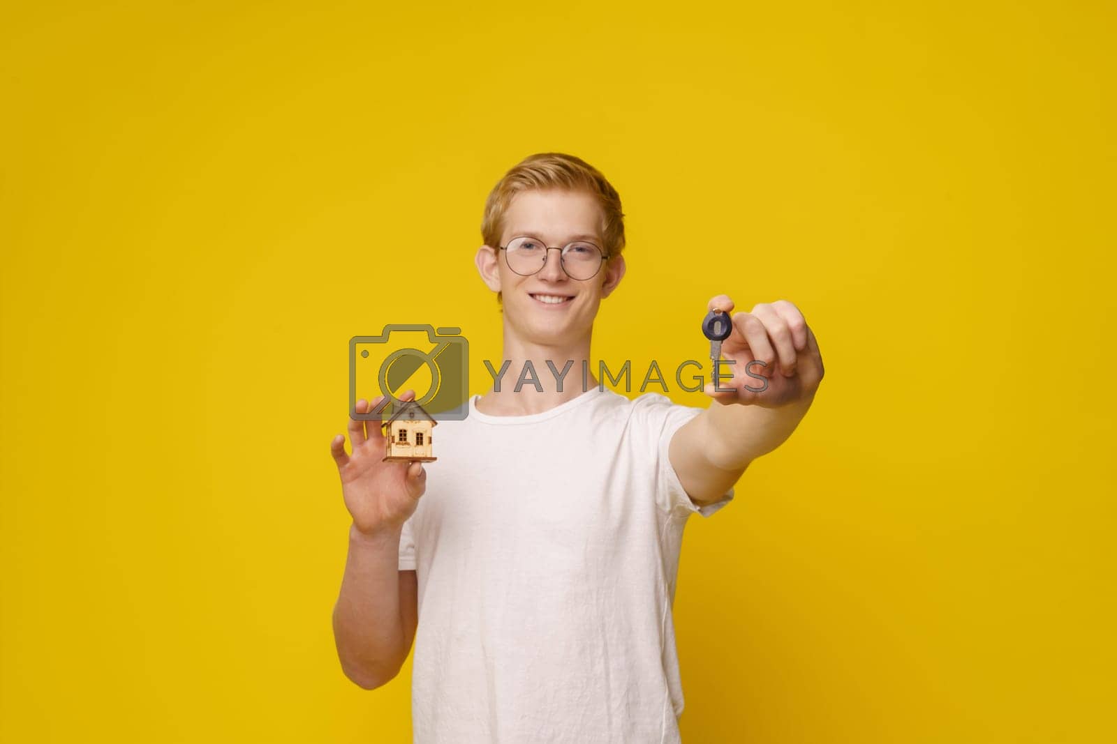 Royalty free image of Young man stands against vibrant yellow background, holding set of keys and model house, symbolizing home ownership and the real estate industry. Focus on hand with key. by LipikStockMedia