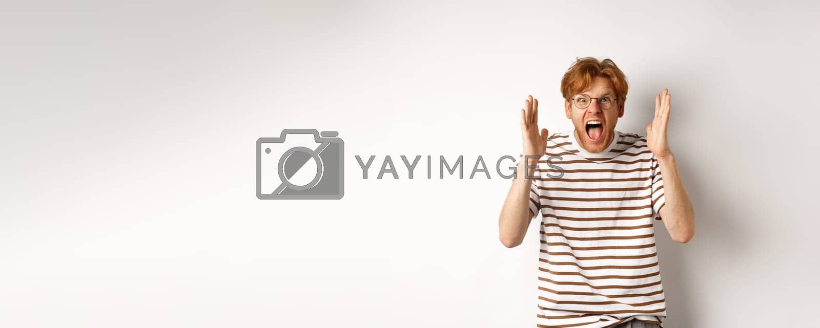 Royalty free image of Angry young man with red hair shouting at camera, screaming and looking outraged, shaking hands, standing over white background by Benzoix