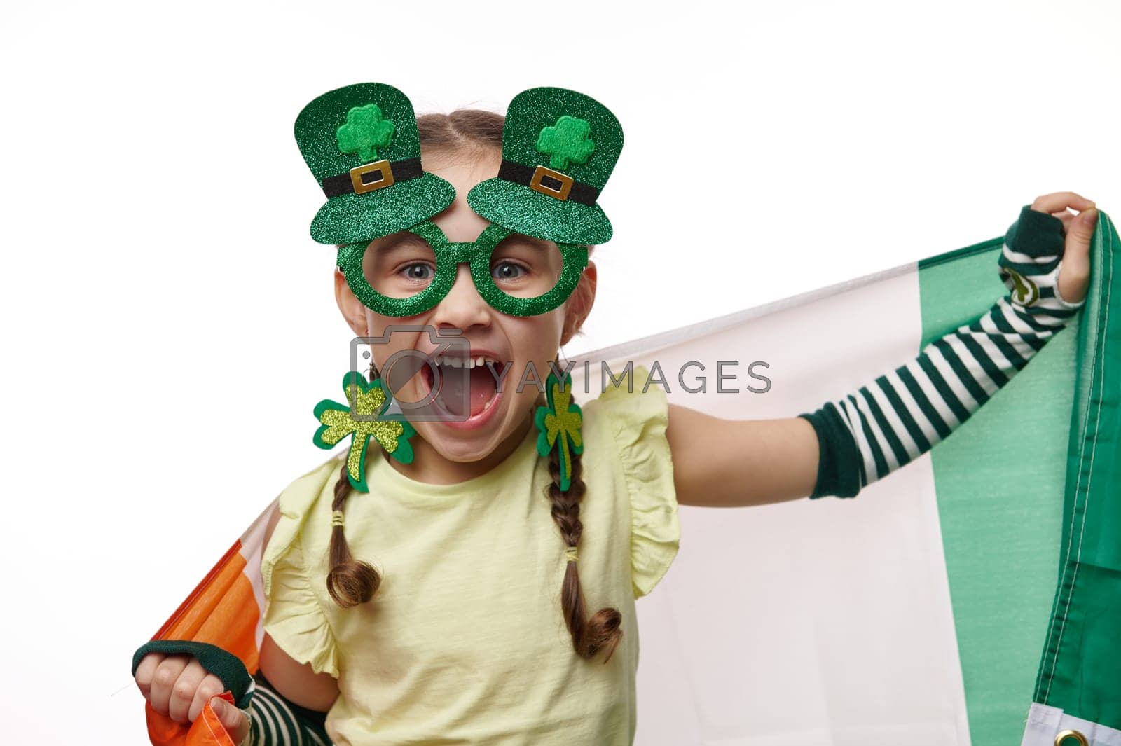 Royalty free image of Irish little girl wearing stylish carnival eyewear, dressed as Leprechaun, carrying Ireland flag, on white background by artgf