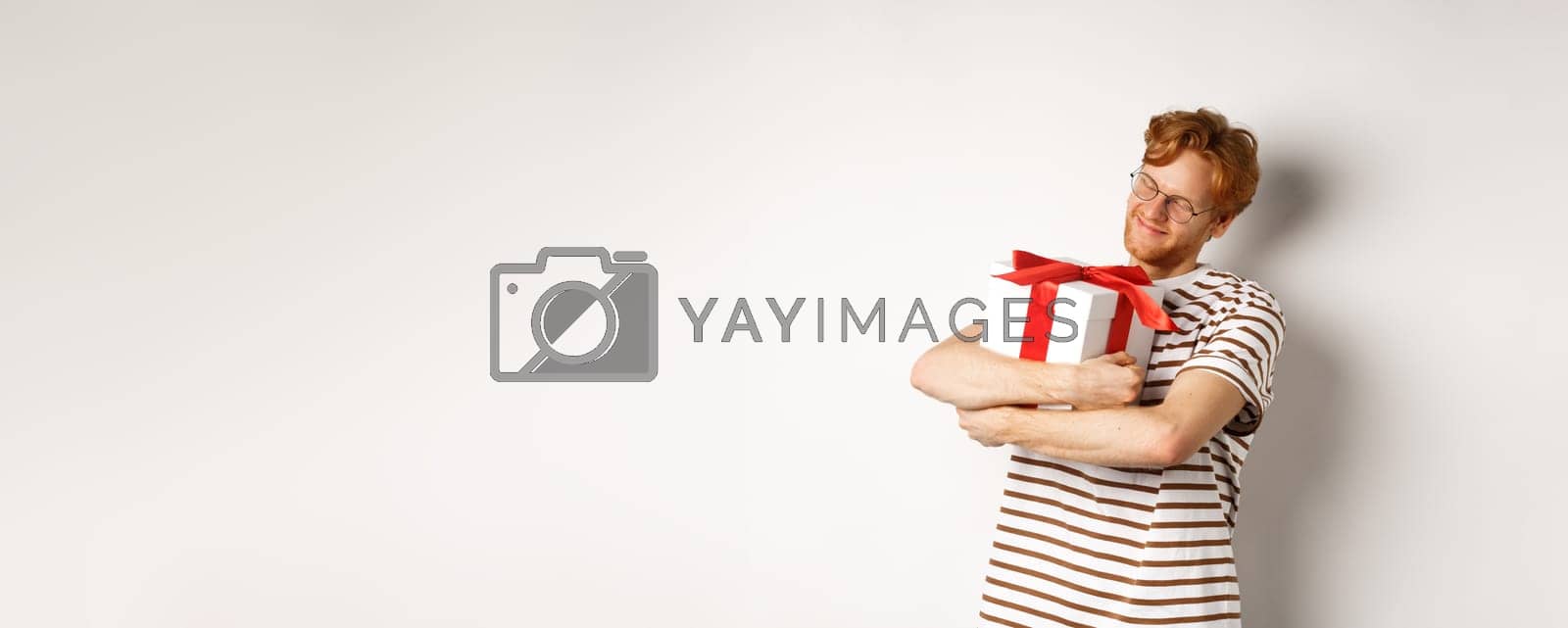 Royalty free image of Valentines day and holidays concept. Happy redhead man hugging his gift box and smiling delighted, standing over white background by Benzoix