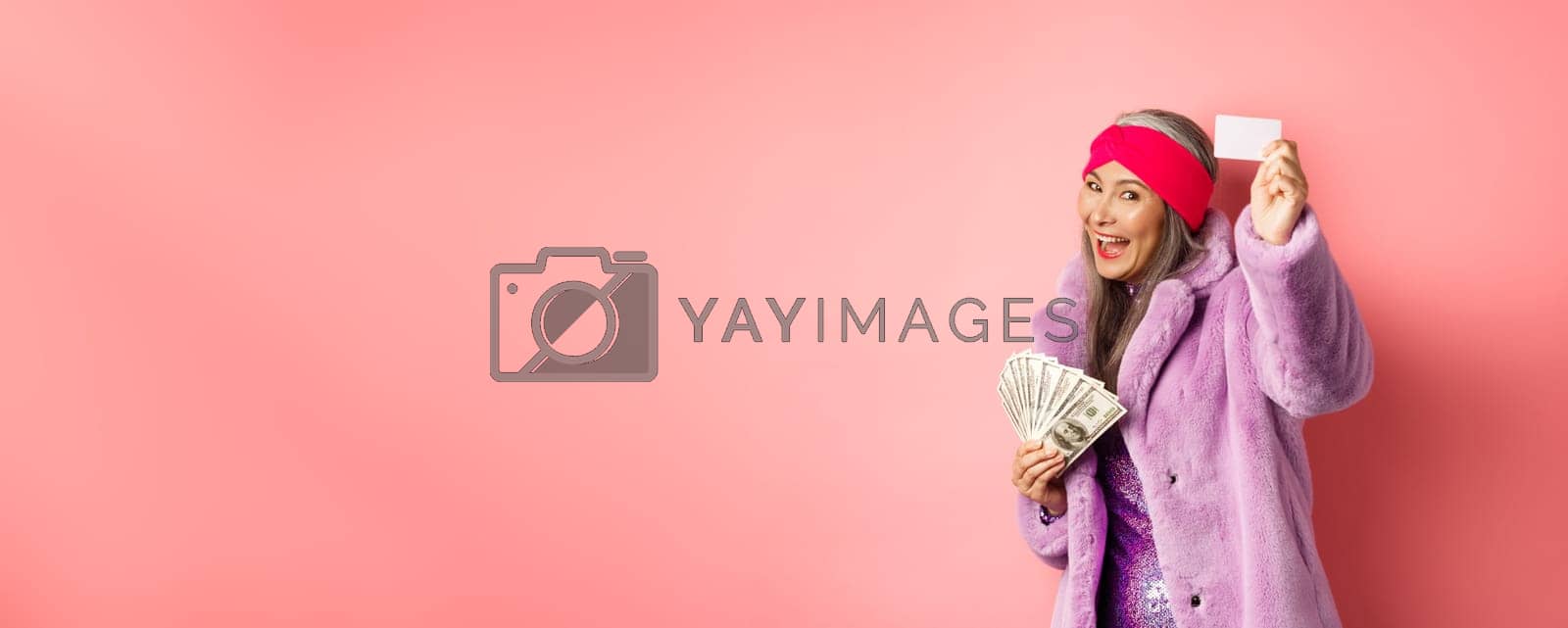 Royalty free image of Shopping and fashion concept. Fashionable funky asian woman showing plastic credit card, paying contactless, holding money in other hand, pink background by Benzoix