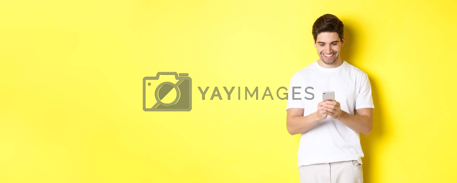 Royalty free image of Young man reading text message on smartphone, looking at mobile phone screen and smiling, standing in white t-shirt against yellow background by Benzoix