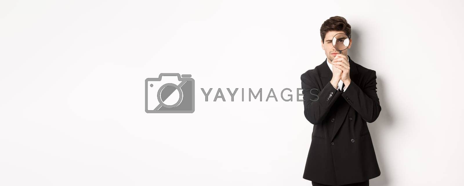 Royalty free image of Image of serious businessman in black trendy suit, looking through magnifying glass, looking for employees, standing against white background by Benzoix