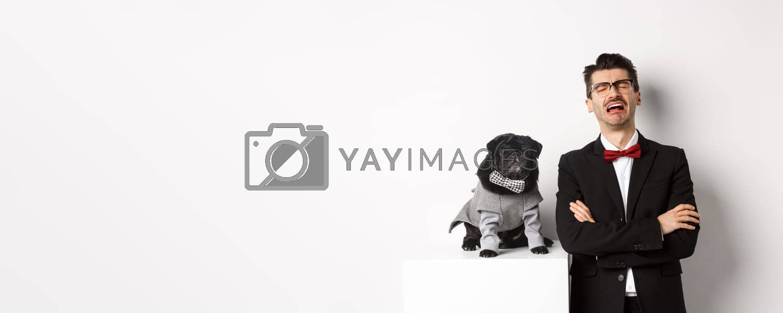 Royalty free image of Animals, party and celebration concept. Sad dog owner crying, wearing suit, standing near cute black pug in costume, standing over white background by Benzoix