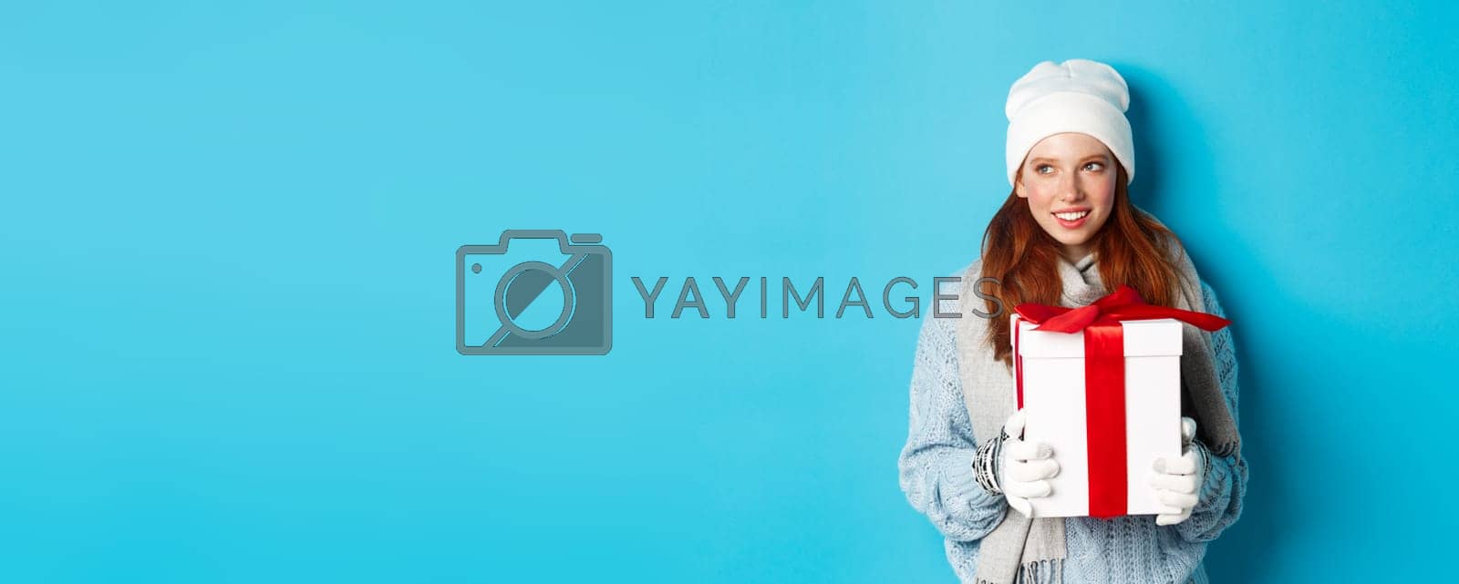 Royalty free image of Winter holidays and Christmas sales concept. Thoughtful redhead woman standing in white beanie and gloves, holding holiday gift and looking left, thinking, standing over blue background by Benzoix