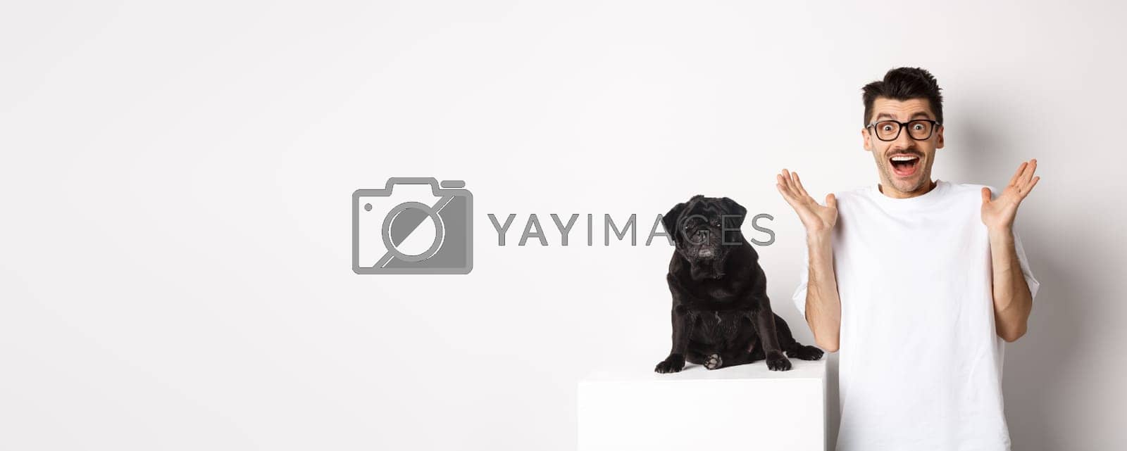Royalty free image of Cheerful young man in glasses standing with his pet, rejoicing and staring at camera amused, hear great news, standing with pug over white background by Benzoix
