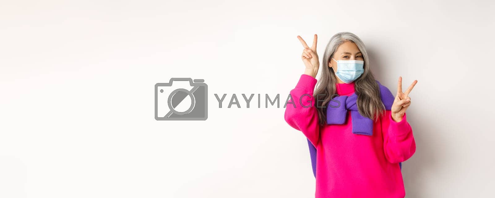Royalty free image of Covid, pandemic and social distancing concept. Cheerful and stylish asian senior woman wearing medical mask and showing peace signs, standing over white background by Benzoix