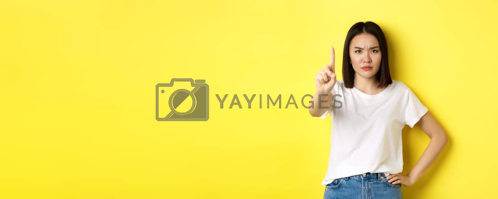 Royalty free image of Confident and serious woman tell no, showing extended finger to stop and prohibit something bad, frowning and looking at camera self-assured, standing over yellow background by Benzoix