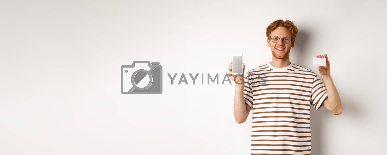 Royalty free image of Shopping and finance concept. Young man showing blank mobile screen and plastic credit card, smiling at camera, white background by Benzoix