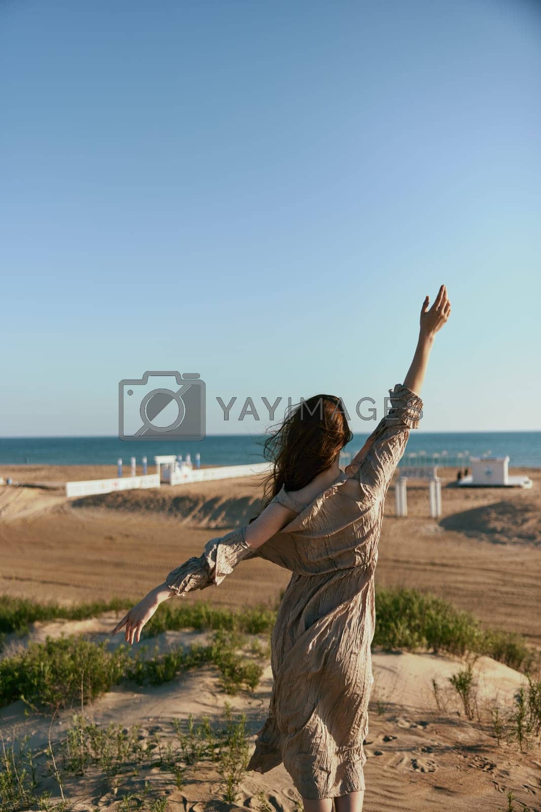 Royalty free image of a woman in a beige dress posing standing on the seashore with her back to the camera with her arms raised high enjoying the view by Vichizh