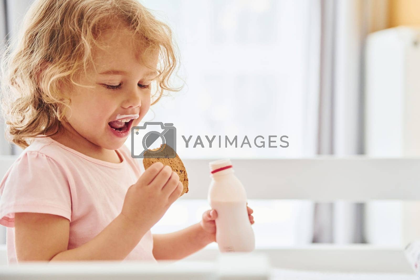 Royalty free image of Sitting on the bed, eating cookies and drinking milk. Cute little girl in casual clothes is indoors at home at daytime by Standret