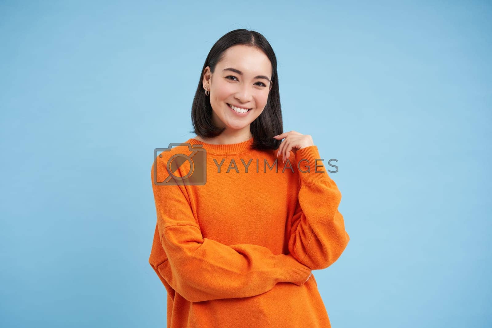 Royalty free image of Cute smiling asian woman, student in orange shirt, looks cute against blue background. Student and lifestyle concept by Benzoix