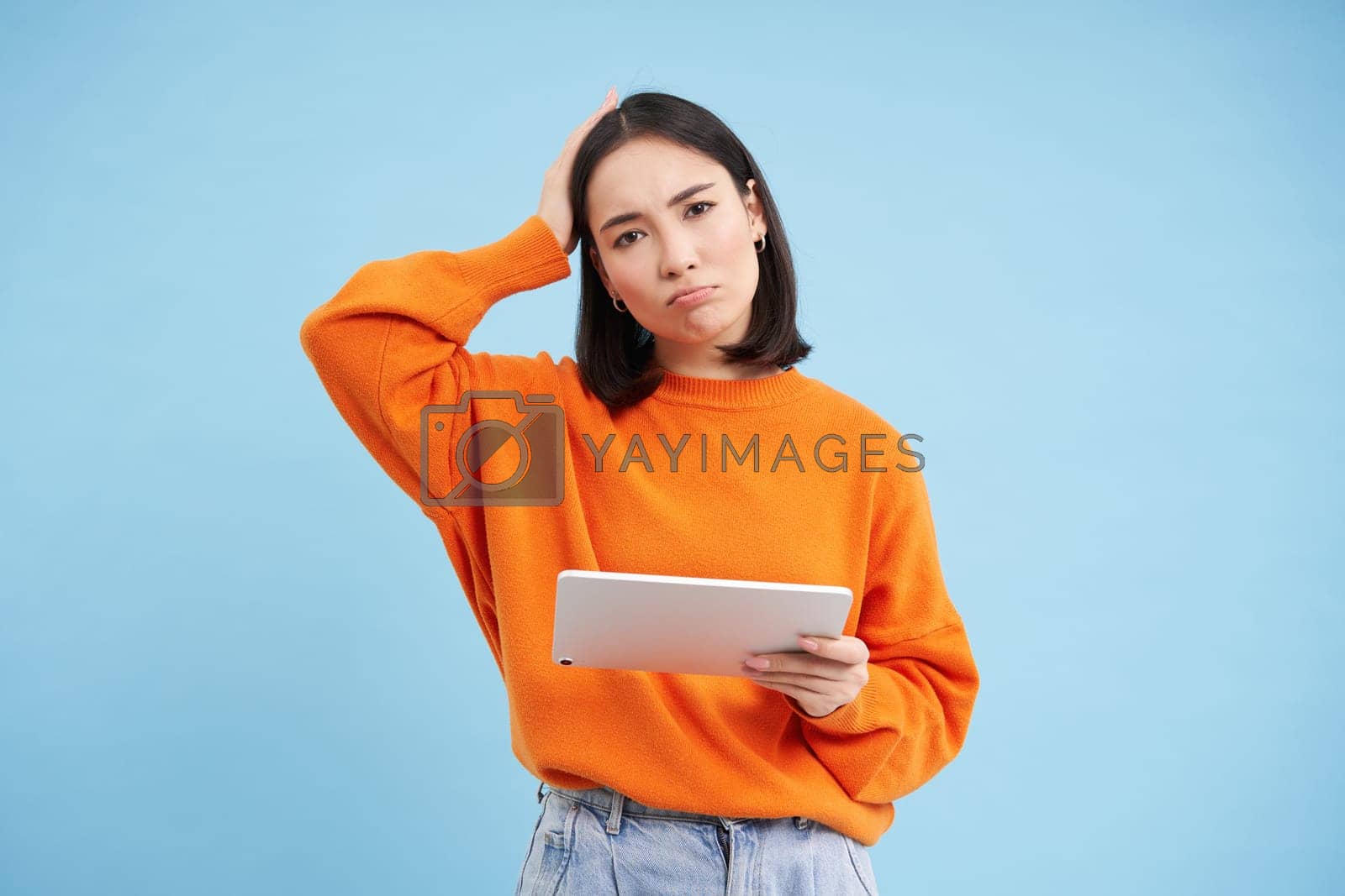 Royalty free image of Woman with digital tablet, looks confused, cant understand smth on her gadget, stands puzzled against blue background by Benzoix