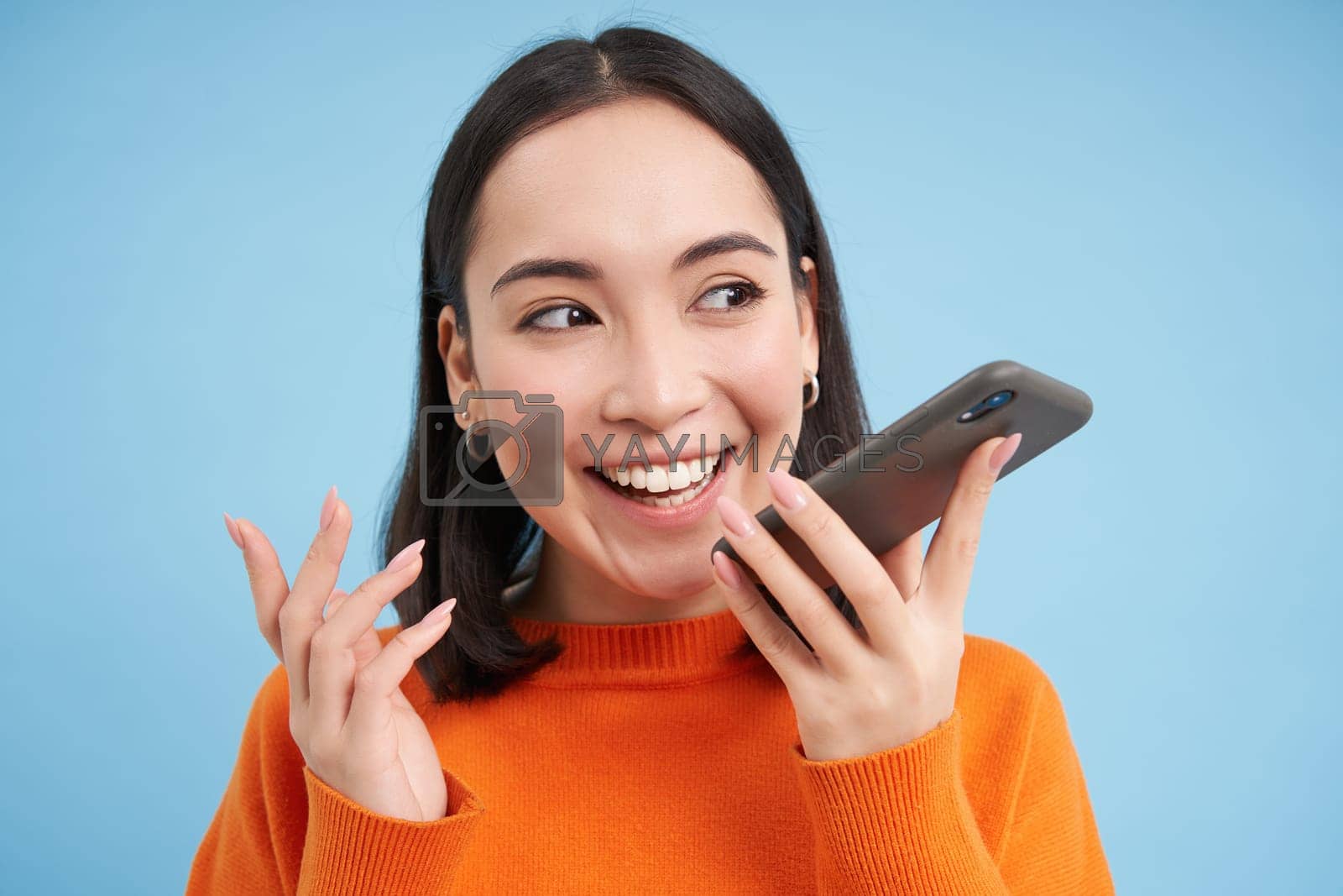 Royalty free image of Portrait of asian woman records voice message, talks on speakerphone, holds mobile phone near mouth while speaking, translates her speech on app, blue background by Benzoix