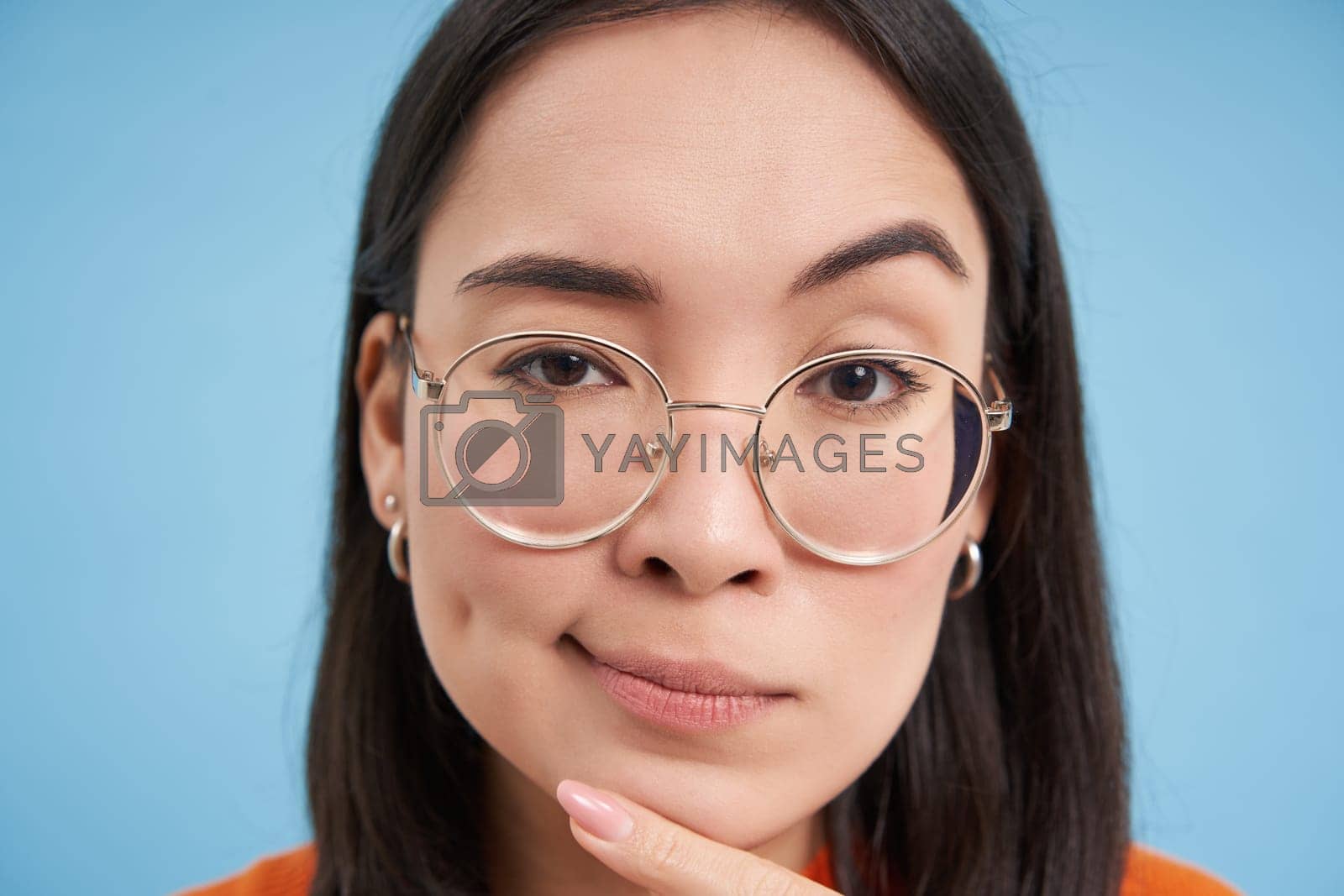 Royalty free image of Close up portrait of asian woman looks intrigued, wears glasses, squints thoughtful, thinking, making assumption, standing over blue background by Benzoix