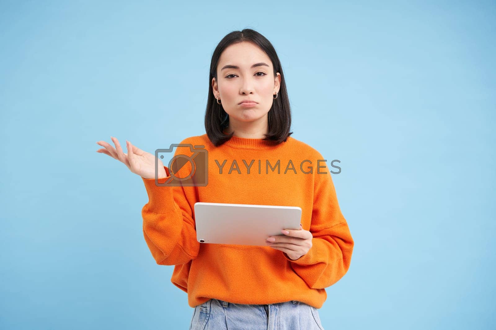 Royalty free image of Portrait of unsure girl, asian woman with digital tablet, shrugging shoulders and looks complicated, standing over blue background by Benzoix