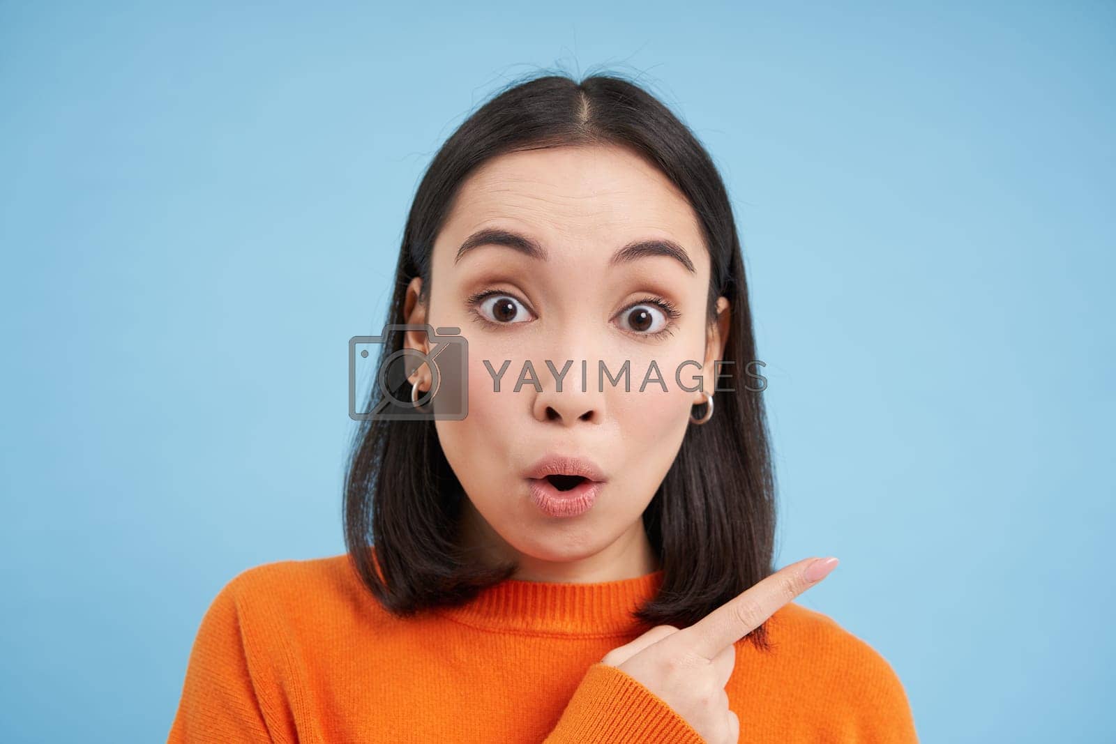 Royalty free image of Close up of surprised asian girl, pointing finger right, showing advertisement with curious, intrigued face, standing over blue background by Benzoix