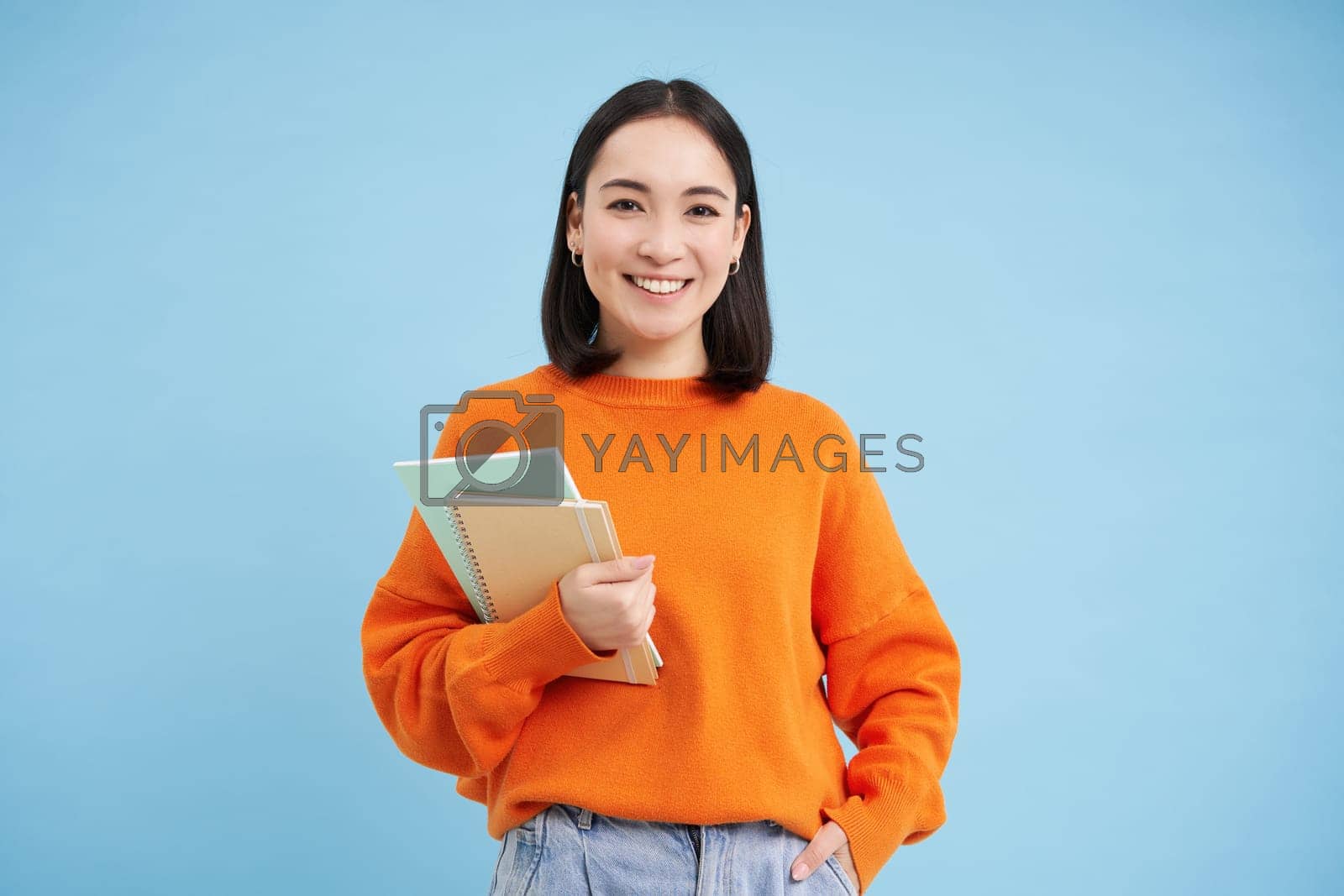 Royalty free image of Confident smiling korean woman with notebooks, University student posing against blue background by Benzoix