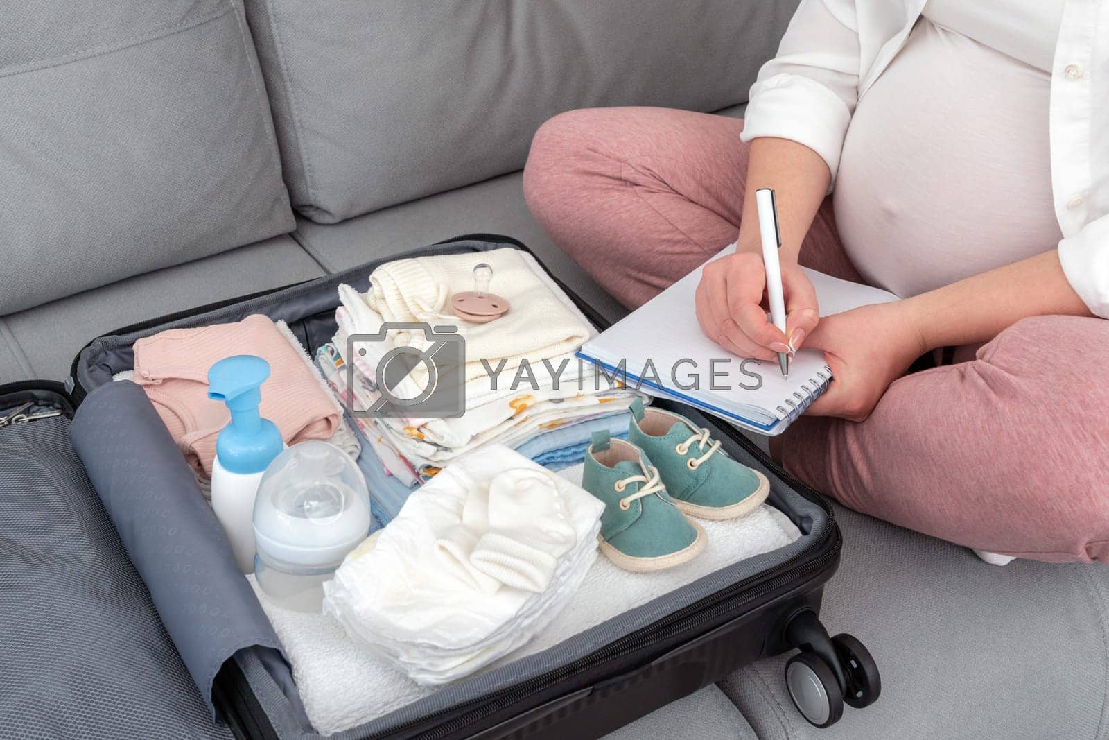 Royalty free image of Pregnant woman writing packing list for maternity hospital sitting on a couch next to suitcase by sdf_qwe