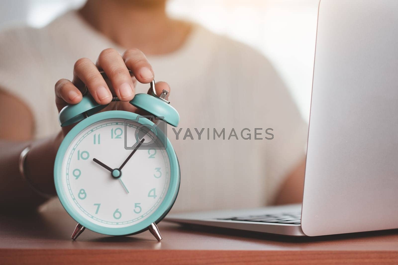 Royalty free image of Asian woman's hand reaching for an alarm clock to check the time on desk with a computer laptop by iamnoonmai