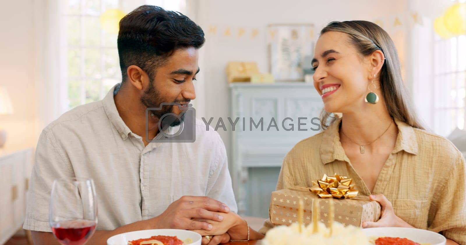 Royalty free image of Interracial couple, gift and celebrate birthday being happy, kiss and smile in home at table with cake. Love, man and woman being content, romantic and present being cheerful celebration together. by YuriArcurs