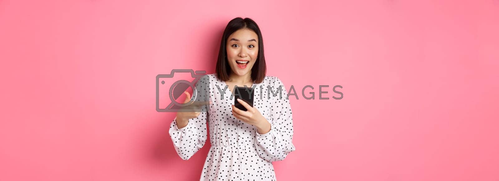 Royalty free image of Online shopping and beauty concept. Amazed and happy asian woman pointing at mobile phone, talking about internet promo offer or app, standing over pink background by Benzoix